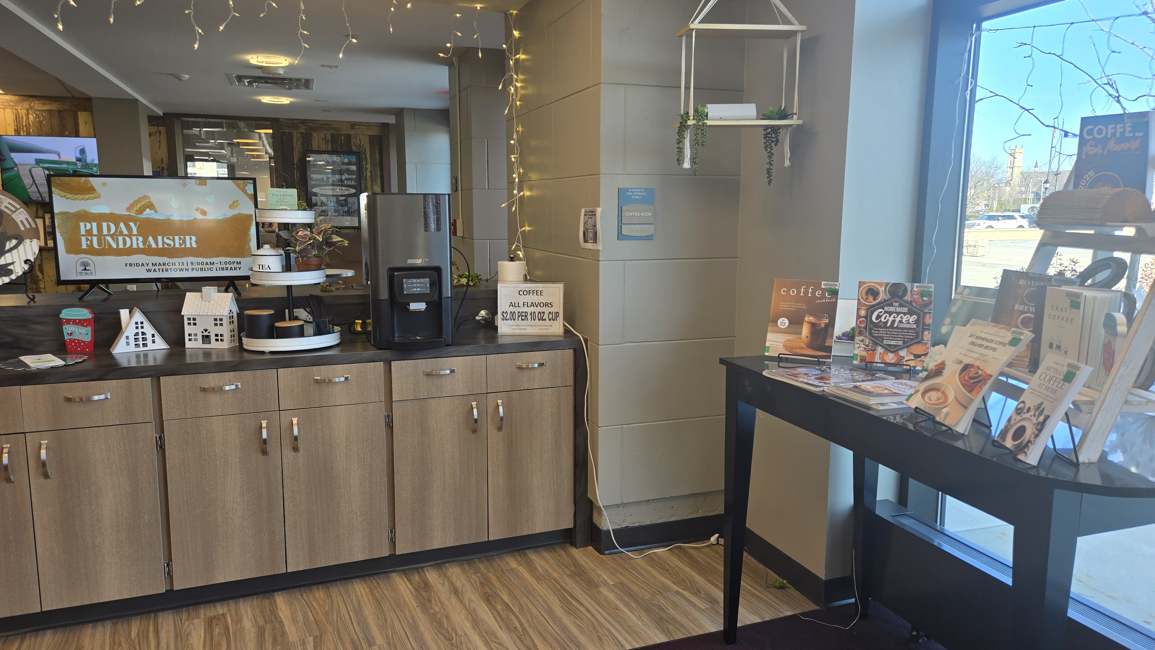 Coffee maker on counter with cupboards below. Flat screen tv on counter also, advertising an upcoming program. Table to the right, in front of window, displaying books about coffee.