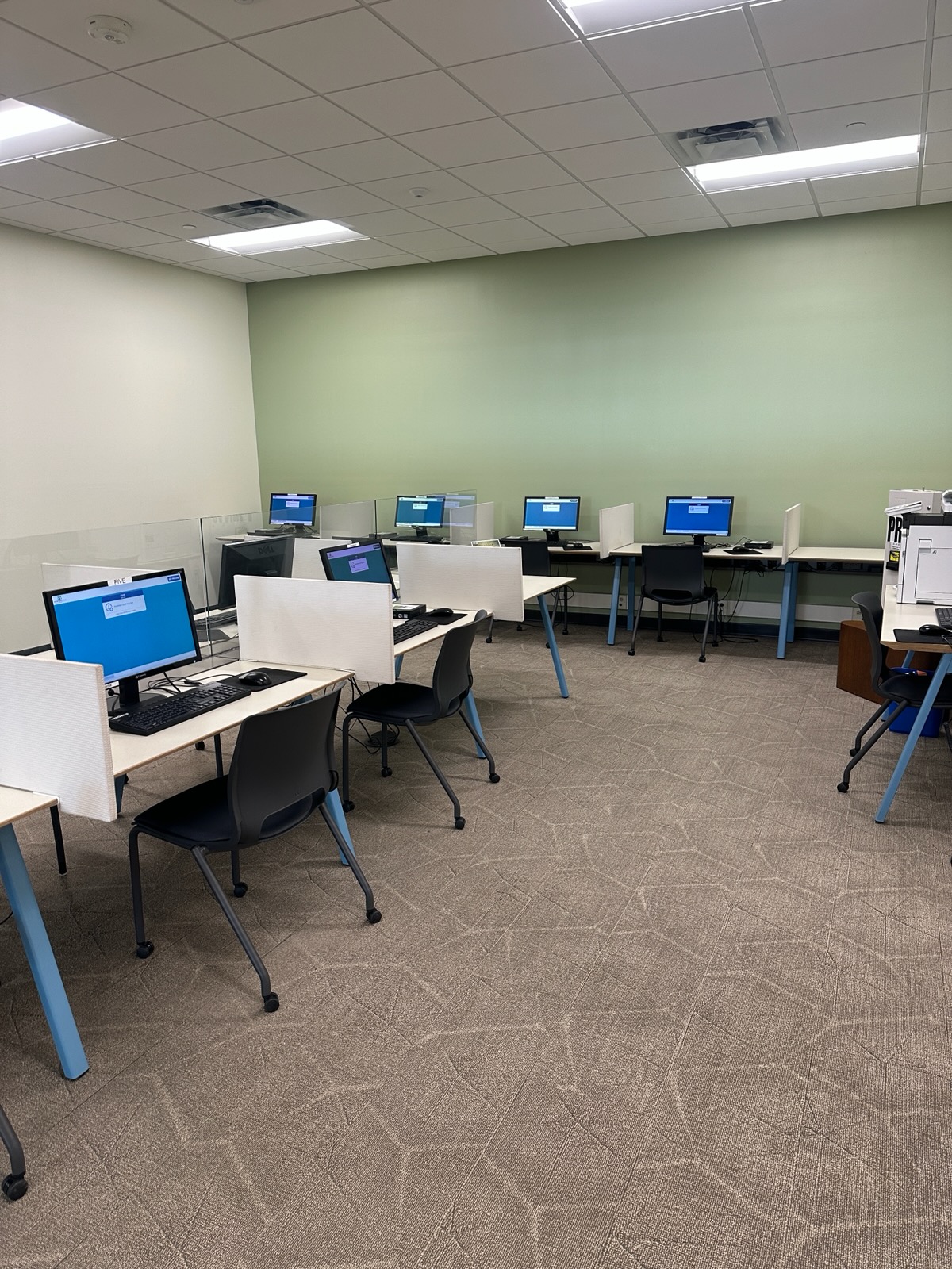 Computer lab with 7 computers set on tables, with black chairs on wheels at each station. Light green wall in the back.