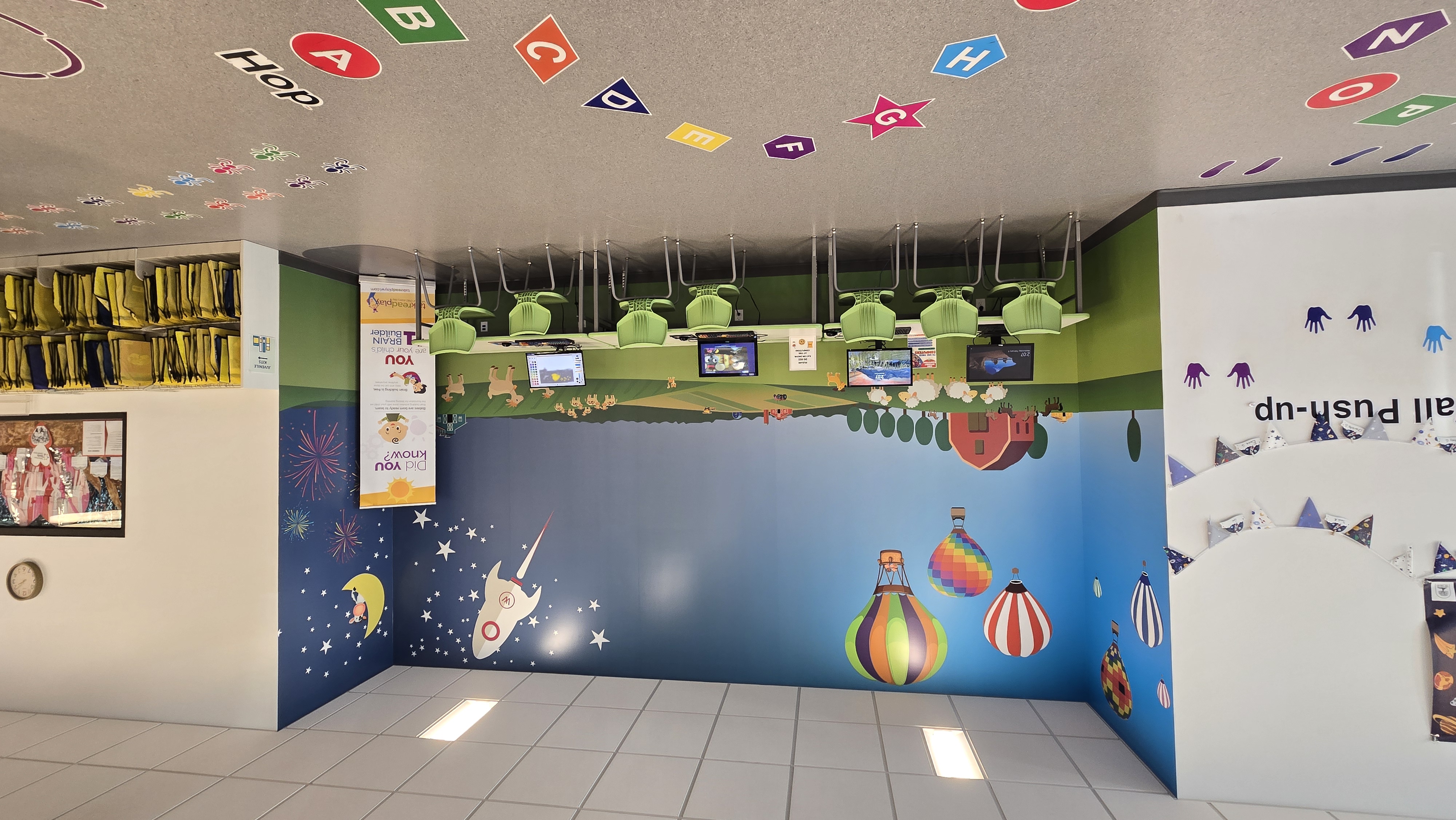 Set of four computers along a wall with farm scene wallpaper. Colorful letters on the floor for kids to jump on.