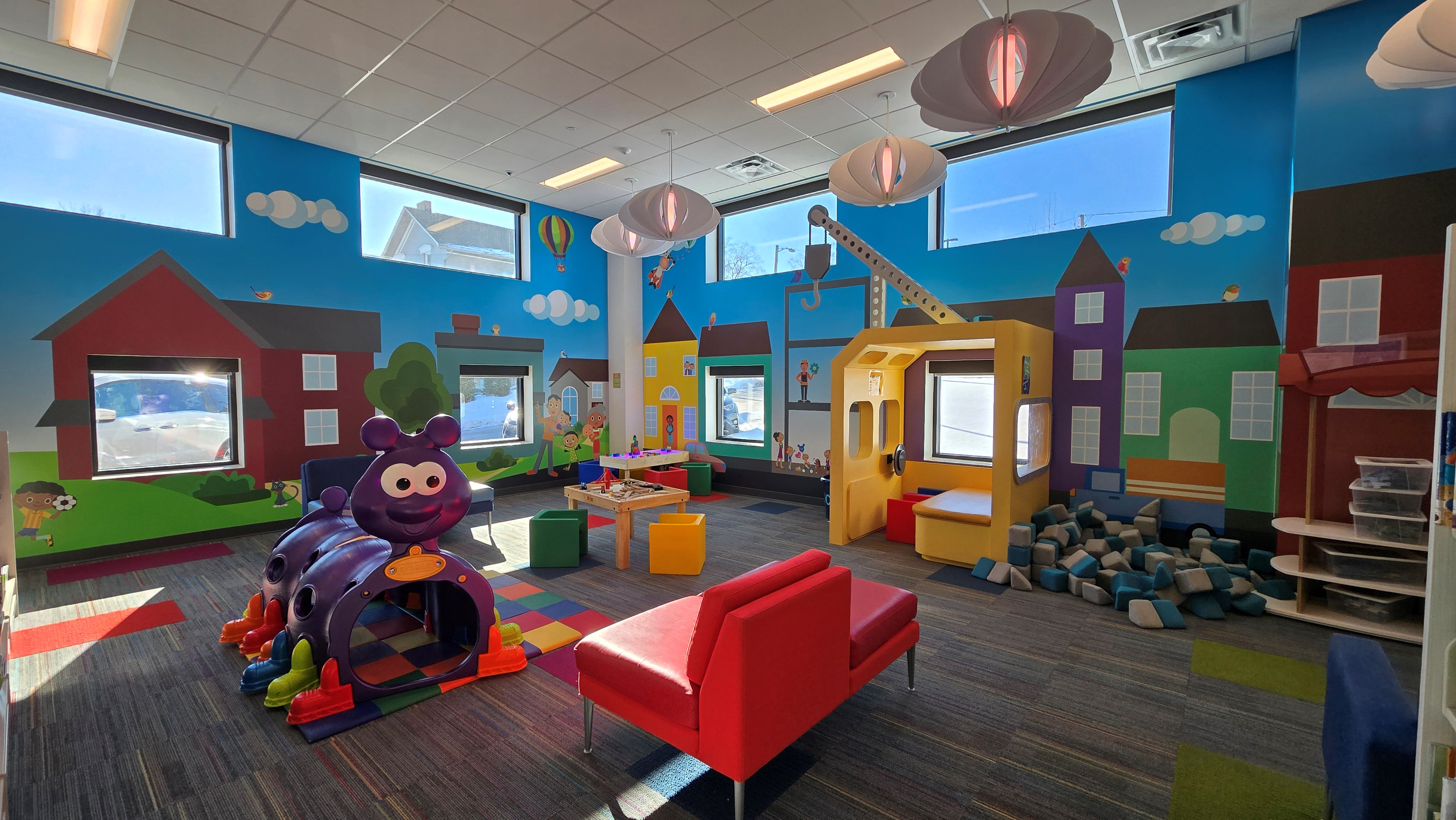 Building blocks with a yellow crane construction vehicle near the wall. Purple caterpillar structure for playing and a red chair for sitting in the center of the shot.