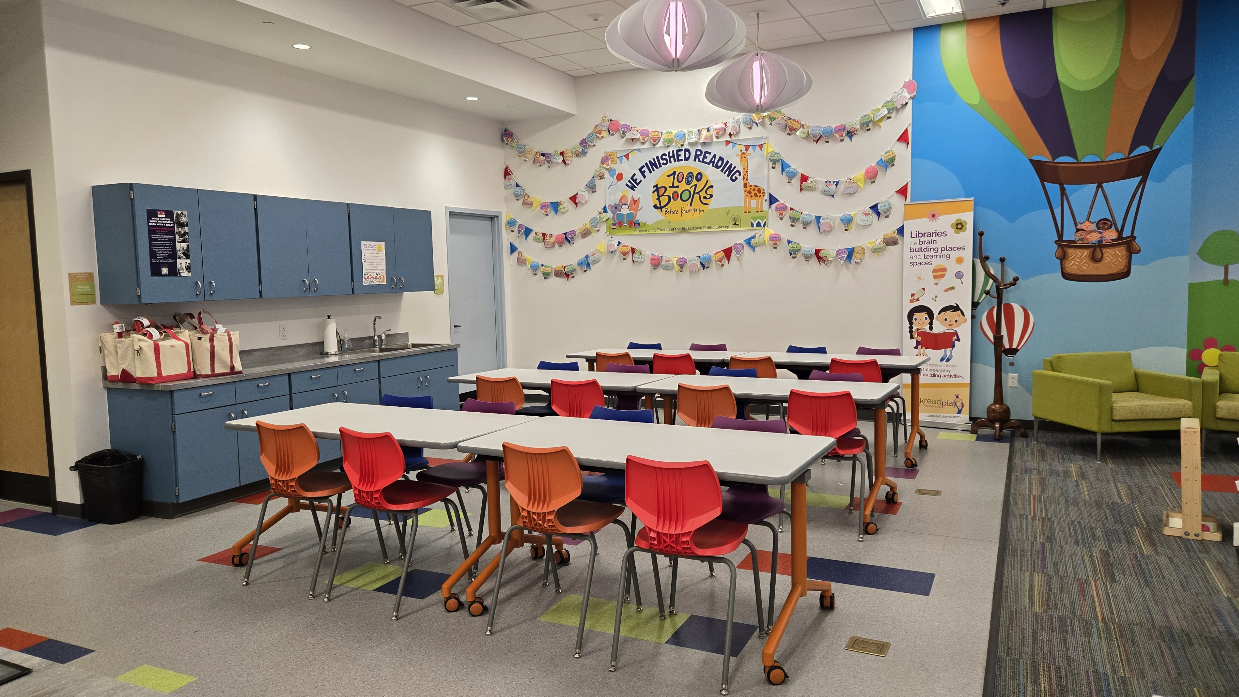 Three long tables with colorful chairs around them. Blue cupboards in the back and colorful banners on the wall in the background.