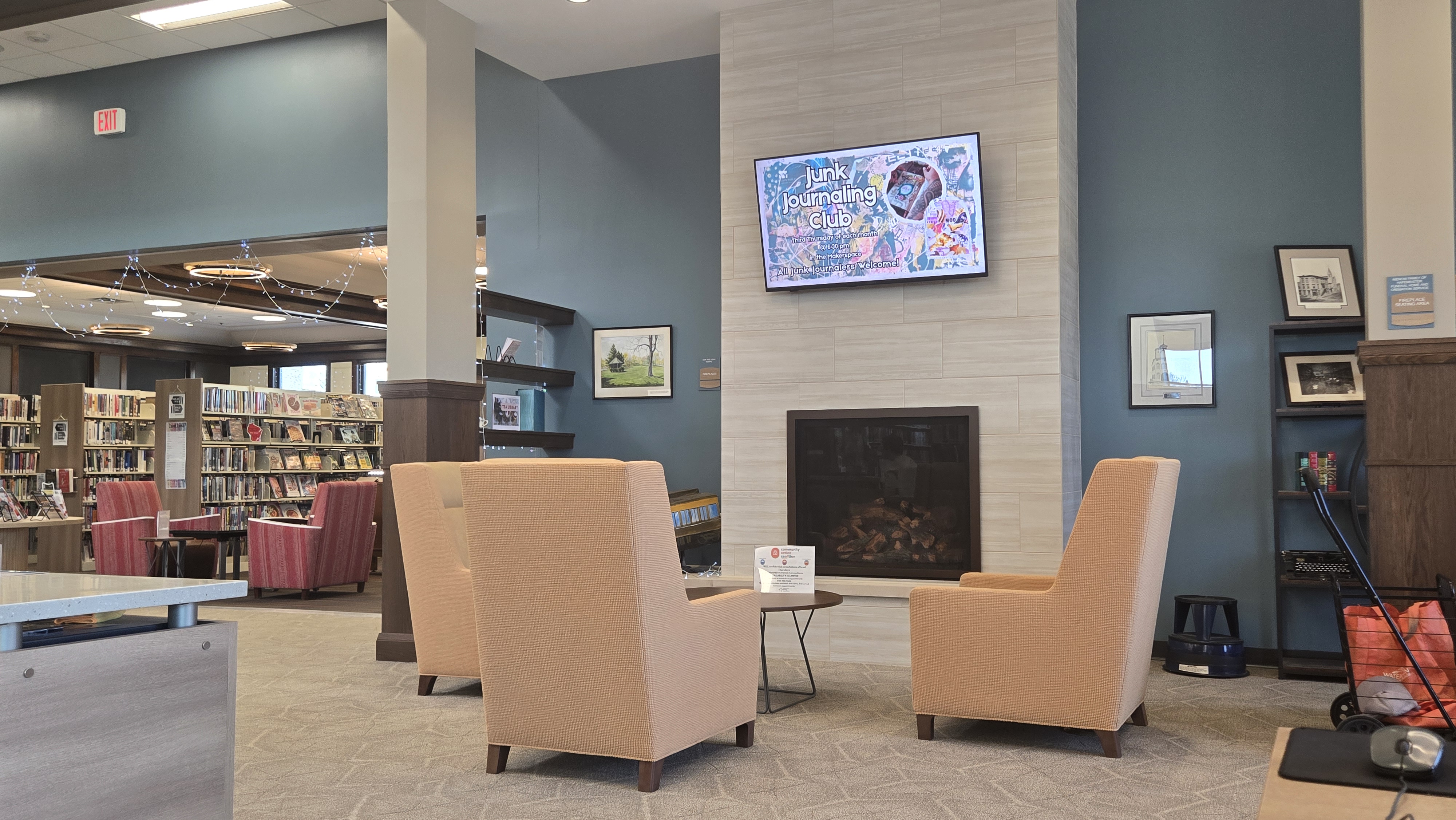 Library scene with three light yellow chairs facing a fireplace. Flat screen tv mounted above the fireplace is showing an advertisement for an upcoming program.