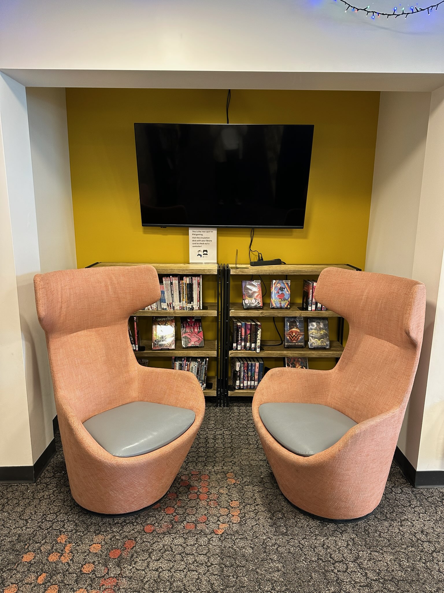 Two modern looking, high-backed orange gaming chairs sitting in front of a book shelf against a mustard yellow wall. A flat screen tv is mounted on the wall.