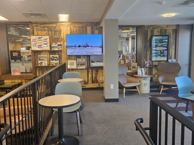 Seating area with small circular tables and chairs. Barnwood wall in background has framed pictures of cows and other farm things, along with a flat screen tv. "Generations of Pride" written on wall.
