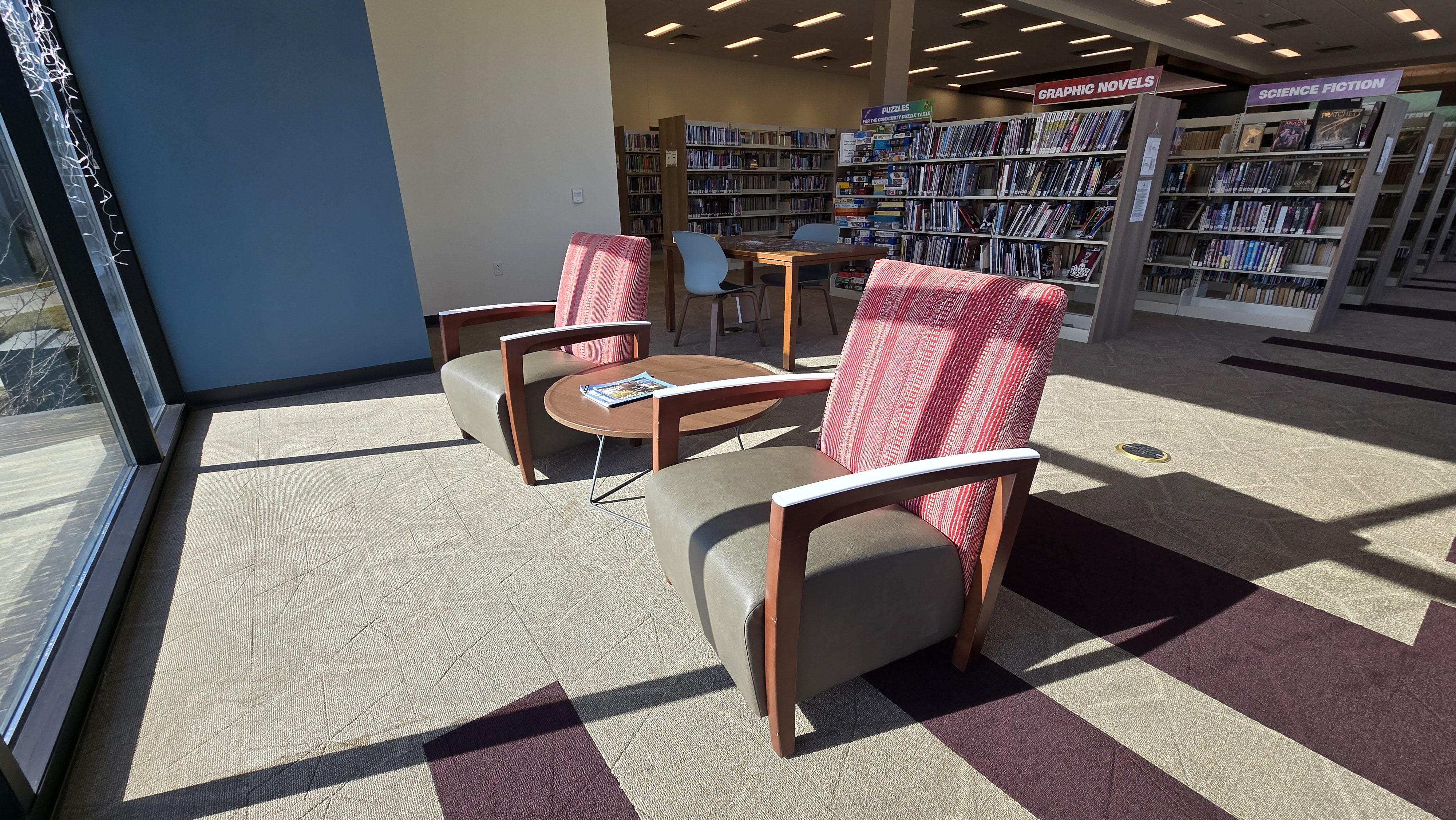 Red, white and brown chairs facing a window in the sun. Large, round coffee table between the two chairs.
