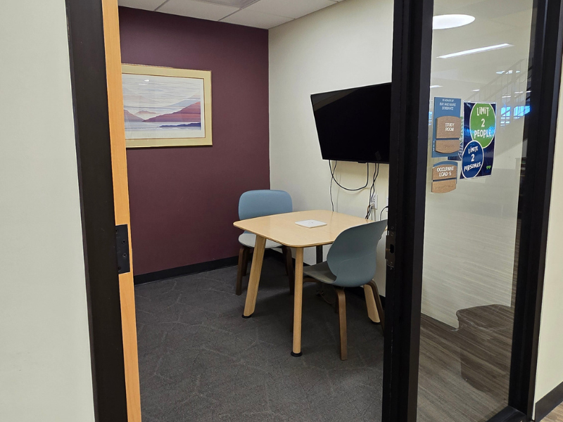 Small room containing table with two chairs. Purple wall in the background with a painting on it. Flat screen tv mounted on wall to the right.
